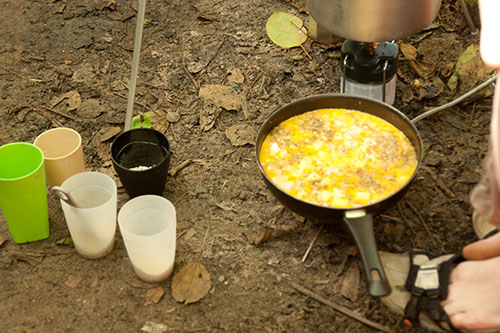 Cozinha do acampamaneto preparando um omelete, exibindo um pé no canto da foto